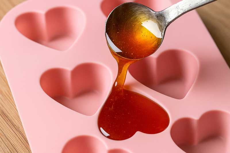 Hot, amber-colored candy syrup being poured directly into a heart-shaped silicone mold without any issues.